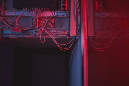 A imagem mostra uma sala técnica ou um centro de dados, onde fios e cabos estão organizados de maneira desordenada, com uma iluminação vermelha suave. Os fios estão pendurados de forma caótica, conectados a equipamentos na parte superior da imagem, e a iluminação cria um ambiente quase sombrio e tecnológico. O fundo da imagem é escuro, sugerindo que o foco está nos cabos e nos dispositivos que os conectam.