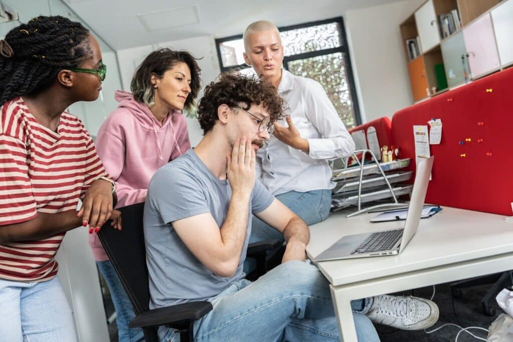 A imagem mostra um grupo de quatro pessoas em um ambiente de escritório. Três delas estão em pé ao lado de uma mesa, observando atentamente uma pessoa sentada, que parece estar preocupada enquanto olha para o computador à sua frente. A pessoa sentada está com a mão na cabeça, parecendo estar pensando ou tentando entender algo. As outras três pessoas, que estão de pé, parecem estar discutindo ou ajudando com o problema. O ambiente é moderno, com móveis e equipamentos típicos de um escritório.