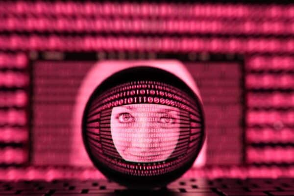 A imagem é uma composição digital com um fundo de padrões de números binários (0s e 1s). No centro da imagem, há um círculo com o rosto de uma mulher visível dentro, e os números binários parecem formar uma grade ao redor dela, sugerindo um tema relacionado à tecnologia e à digitalização. A cena tem um tom de cor rosa e cria uma sensação futurista, com um foco visual no rosto da mulher no centro, que está parcialmente refletido.