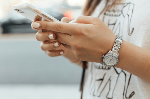 A imagem mostra uma pessoa segurando um smartphone com uma mão, enquanto a outra mão está visivelmente segurando um relógio de pulso prateado. A pessoa parece estar usando o celular, com as mãos em destaque, e as unhas estão pintadas de branco. A camisa que ela está usando tem um desenho de elefante. O foco da imagem está nas mãos e no relógio, com o fundo desfocado, sugerindo um ambiente externo.