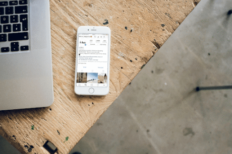 A imagem mostra um ambiente de trabalho simples e moderno. À esquerda, há um laptop aberto sobre uma mesa de madeira, com parte da tela visível. Ao lado, em cima da mesa, está um celular, com a tela exibindo a interface do Instagram, possivelmente mostrando o perfil de um usuário. A mesa tem um acabamento rústico, e o fundo sugere que o local pode ser um escritório ou espaço de trabalho. A imagem captura uma sensação de produtividade e conectividade.