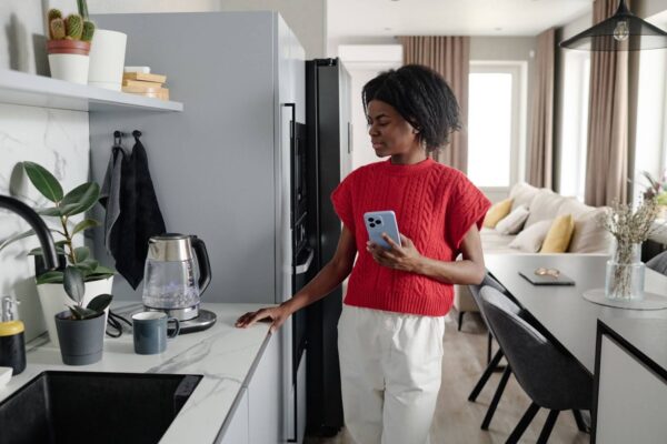 A imagem mostra uma mulher em uma cozinha moderna, segurando um celular enquanto observa uma chaleira elétrica que está sendo ligada. Ela está usando um suéter vermelho e calças brancas, com uma expressão pensativa. A cozinha é iluminada por luz natural, e o ambiente é clean, com móveis modernos e plantas decorativas na bancada. O espaço ao fundo parece acolhedor e bem organizado, com uma sensação de conforto e tranquilidade.