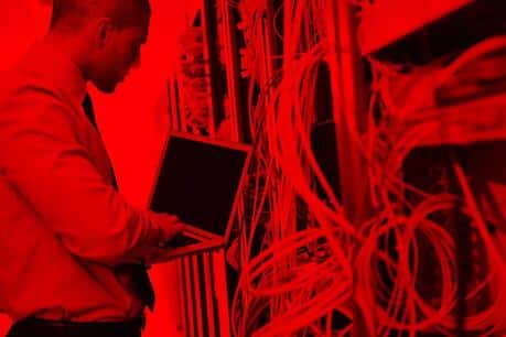 Um homem em pé, usando terno claro, está diante de um rack de servidores com vários cabos conectados. Ele segura um notebook aberto e parece realizar manutenção ou configuração. Toda a cena está iluminada com uma forte luz vermelha.