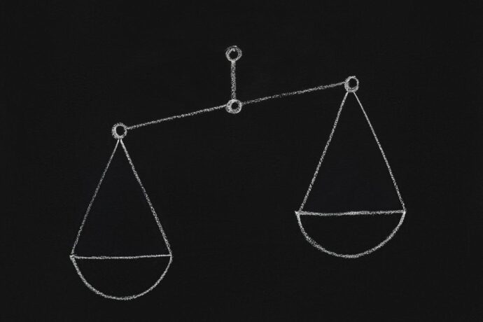 A imagem mostra um desenho de uma balança feita com giz, com o fundo preto. As duas tigelas da balança estão desenhadas de forma simétrica, penduradas em um eixo central. O desenho é simples, mas transmite a ideia de equilíbrio e justiça, com uma linha fina conectando as duas partes da balança.