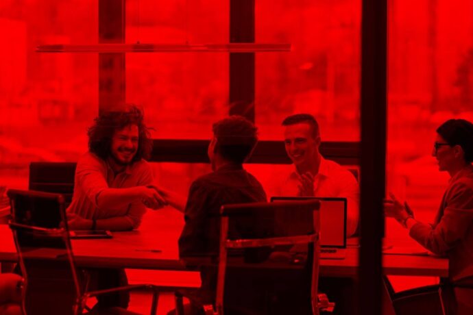 A imagem retrata um grupo de quatro pessoas em uma reunião, sentados em torno de uma mesa em um ambiente com uma iluminação vermelha intensa, o que dá à cena uma tonalidade quente e enigmática. Eles estão sorrindo e interagindo, com duas pessoas apertando as mãos no centro da mesa. As outras duas estão sentadas, observando, uma delas aplaudindo. O ambiente é moderno, com móveis de escritório e laptops visíveis.
