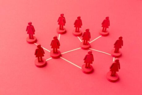 A imagem mostra figuras em forma de mulheres, feitas de peças de madeira ou plástico, dispostas em uma rede conectada por linhas brancas. Cada figura está em um círculo e as linhas unem as diferentes figuras, sugerindo uma rede ou conexão entre elas. O fundo é de uma cor vermelha sólida, o que dá um tom forte e vibrante à imagem.