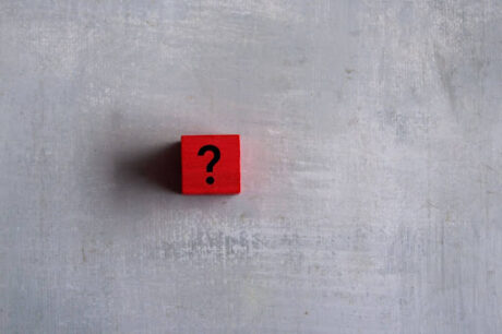 A imagem mostra um bloco de cubo vermelho com um símbolo de interrogação preto impresso em um dos lados. O cubo está posicionado de forma que a interrogação seja claramente visível, sugerindo que há algo que precisa ser resolvido ou explicado. O fundo é claro e neutro, o que faz o cubo se destacar visualmente.