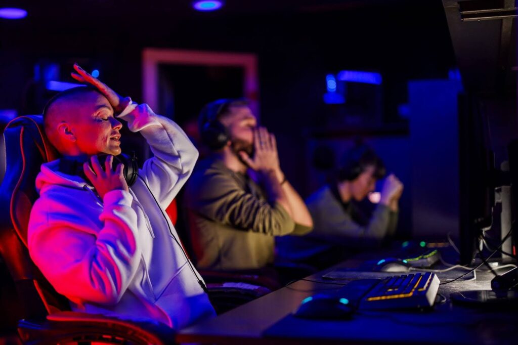 A imagem mostra um grupo de três pessoas jogando videogame em um ambiente de sala escura, com luzes coloridas que iluminam suavemente o espaço. Duas pessoas estão visivelmente frustradas: uma delas tem a mão na cabeça, expressando desespero ou tensão, enquanto a outra está com as mãos juntas, como se estivesse rezando ou esperando algo. A terceira pessoa, ao fundo, também parece estar concentrada no jogo. O ambiente é moderno, com cadeiras de gamer e equipamentos de última geração, criando uma atmosfera de tensão e emoção.