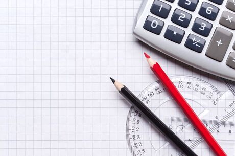 Uma folha quadriculada branca com dois lápis — um preto e um vermelho — posicionados ao lado de um transferidor transparente. No canto superior direito, há parte de uma calculadora cinza.