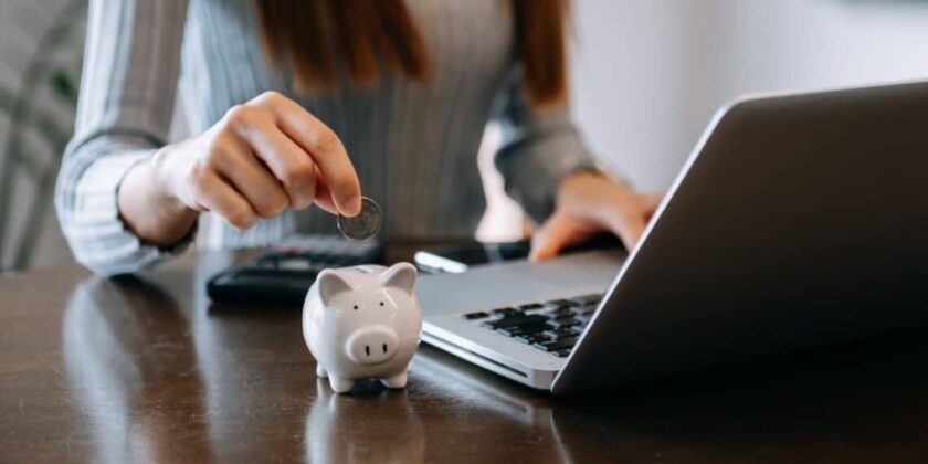 uma mulher sentada em frente ao notebook e colocando uma moeda em um cofrinho