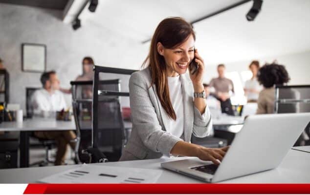 Mulher sorrindo no escritório enquanto fala ao telefone e usa notebook