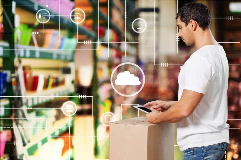 Uma fotografia horizontal de um homem branco, em pé, em um armazém. O homem está vestindo uma camiseta branca e calças jeans. Ele segura um tablet e olha para ele, enquanto se curva sobre uma caixa de papelão que está sobre outra caixa. O fundo da imagem está desfocado, com a iluminação do teto e das prateleiras, e há gráficos de linha branca e ícones sobrepostos à imagem, indicando um conceito de nuvem ou de rede.