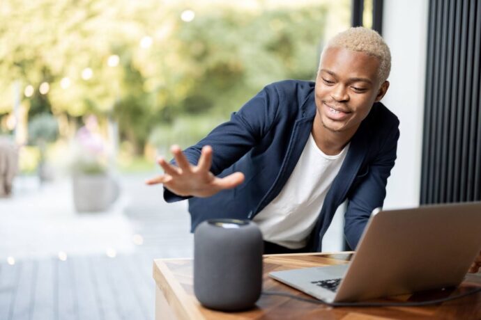 Uma fotografia horizontal de um homem negro, com cabelo descolorido e loiro, vestindo uma camisa branca e um blazer azul-marinho. Ele está em uma área externa, possivelmente uma varanda ou pátio, com a mão direita estendida para um alto-falante cilíndrico e cinza que está em uma mesa de madeira. O homem está sorrindo e olhando para o alto-falante, parecendo interagir com ele. Há um laptop aberto e prateado à sua direita e um fundo com folhagem verde embaçada e luzes de jardim.