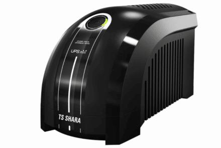Uma fotografia horizontal, com um fundo branco puro, de um no-break pequeno e preto, da marca TS SHARA. O no-break tem um formato curvo e oval, com uma borda cinza na parte de trás. Há um grande botão de ligar e desligar no topo, com uma luz verde acesa. O no-break é pequeno e tem entradas de ar na lateral e na parte de trás.