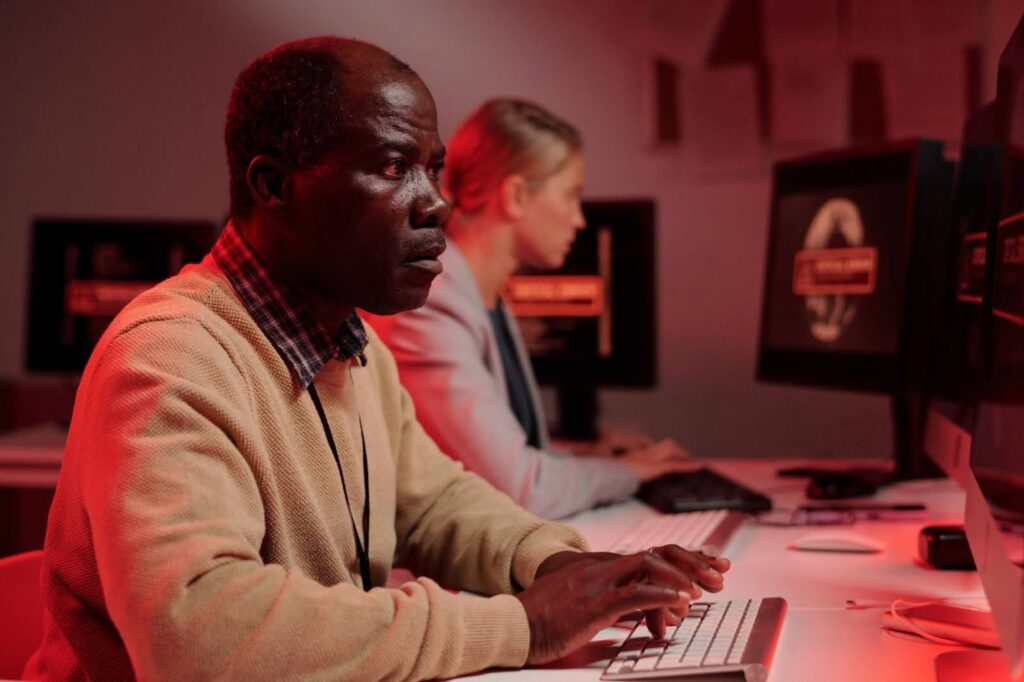 Na imagem, vemos dois indivíduos em um ambiente de trabalho em um escritório com luzes vermelhas suaves. À esquerda, um homem de pele escura, usando uma camisa de malha bege e uma camisa de xadrez por baixo, está concentrado digitando em um teclado. Ele parece estar observando a tela do computador, com uma expressão séria e focada. À direita, uma mulher de pele clara, usando um blazer cinza, está sentada em um computador também, com as mãos em um teclado, voltada para a tela. Ambos estão em frente a monitores, e há papéis na parede atrás deles, sugerindo um ambiente de trabalho intenso. O clima da cena é tenso, com uma iluminação vermelha que dá um tom dramático à imagem.