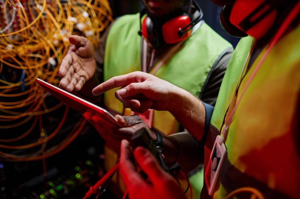 Uma fotografia de perto, horizontal, de dois trabalhadores em uma sala de servidores. Os dois estão vestidos com coletes de segurança verdes e coletes de segurança amarelos, e ambos usam fones de ouvido de segurança grandes e laranja sobre as orelhas. O trabalhador à esquerda é negro e estende a mão com a palma aberta sobre um tablet que o trabalhador à direita, que é branco, está segurando. O dedo indicador do trabalhador à direita está estendido e apontando para a tela do tablet. Um emaranhado de cabos amarelos e laranjas desfocados está em segundo plano, no lado esquerdo. A imagem é dominada por luzes vermelhas, que iluminam os coletes de segurança.