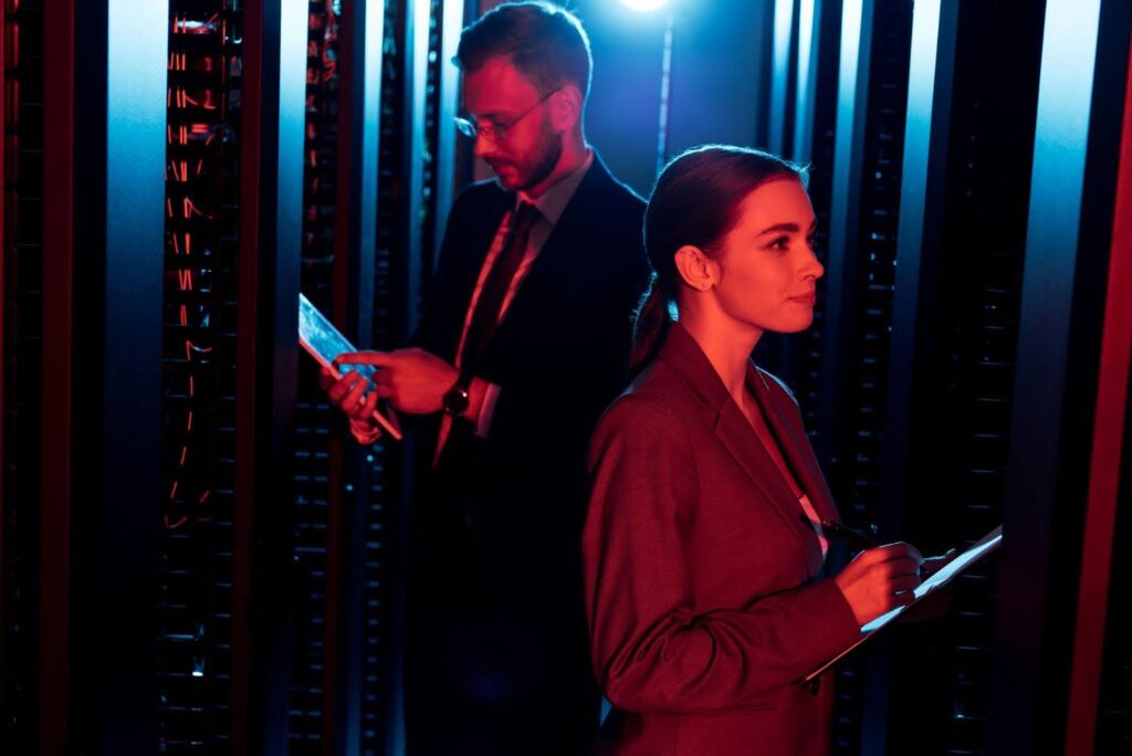 A imagem mostra duas pessoas em um ambiente de data center, entre estantes de servidores. A luz é predominantemente azul e vermelha, criando um efeito dramático. À esquerda, um homem de terno está lendo um documento ou anotando algo, enquanto à direita, uma mulher com cabelo preso, também vestindo um terno, está concentrada em seu caderno, tomando notas. Eles estão em posições opostas, mas no mesmo espaço, em um ambiente de trabalho de tecnologia.