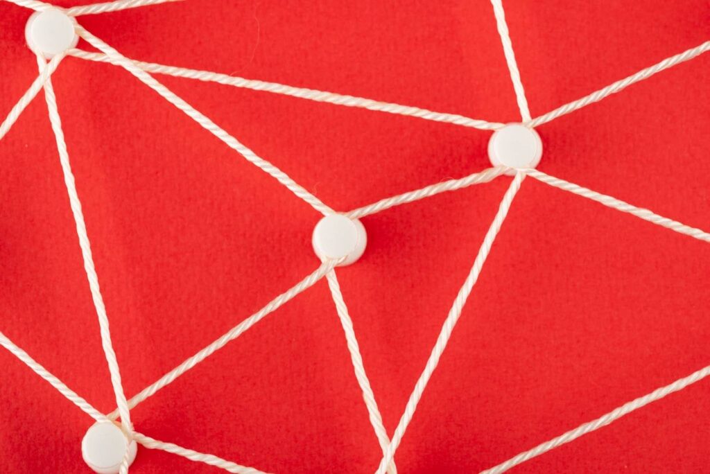 Uma imagem de um fundo vermelho vibrante, com vários pontos circulares brancos unidos por linhas brancas de barbante ou corda. Os pontos brancos são colocados aleatoriamente, e as linhas brancas se cruzam, formando uma variedade de triângulos de tamanhos diferentes, representando uma rede de conexões.