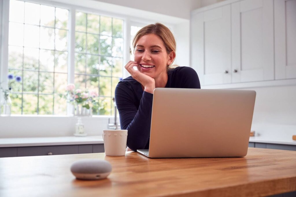 Uma fotografia horizontal de uma mulher loira e branca, sorrindo, em uma cozinha. A mulher está sentada à mesa e olhando para um laptop, que está à sua frente. Há um pequeno alto-falante oval e cinza à esquerda do laptop e uma xícara branca à direita. A mulher está com as mãos juntas, com o rosto apoiado nas mãos, e seu cabelo está amarrado para trás. A janela na parede de trás tem uma grade, e o fundo é verde com árvores e algumas flores cor-de-rosa.