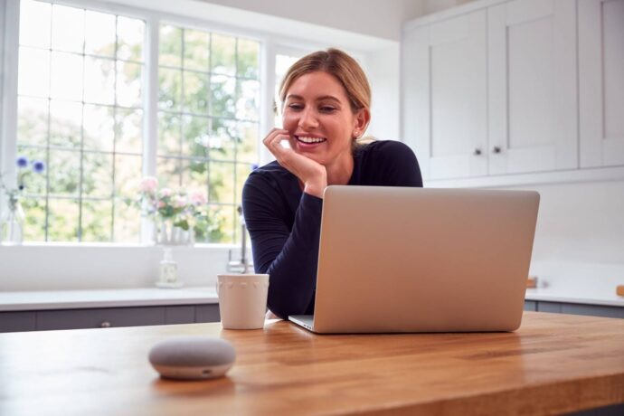 Uma fotografia horizontal de uma mulher loira e branca, sorrindo, em uma cozinha. A mulher está sentada à mesa e olhando para um laptop, que está à sua frente. Há um pequeno alto-falante oval e cinza à esquerda do laptop e uma xícara branca à direita. A mulher está com as mãos juntas, com o rosto apoiado nas mãos, e seu cabelo está amarrado para trás. A janela na parede de trás tem uma grade, e o fundo é verde com árvores e algumas flores cor-de-rosa.