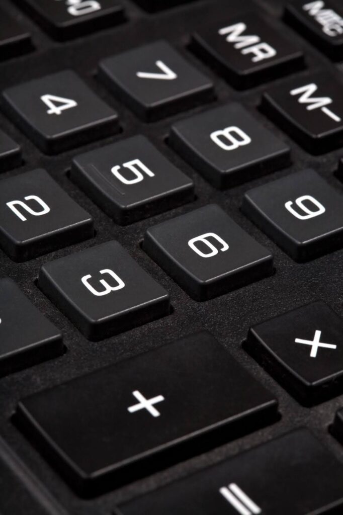 Uma fotografia de perto, horizontal, de um teclado de calculadora ou teclado numérico. O foco está nos botões pretos, com caracteres brancos, que parecem ser de plástico. Os números e os símbolos são grandes e facilmente visíveis, mas o botão de ligar não está focado, parecendo ter uma leve luz verde. O no-break tem um formato arredondado, com uma borda cinza na parte de trás e entradas de ar em ambos os lados.