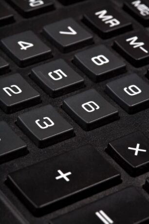 Uma fotografia de perto, horizontal, de um teclado de calculadora ou teclado numérico. O foco está nos botões pretos, com caracteres brancos, que parecem ser de plástico. Os números e os símbolos são grandes e facilmente visíveis, mas o botão de ligar não está focado, parecendo ter uma leve luz verde. O no-break tem um formato arredondado, com uma borda cinza na parte de trás e entradas de ar em ambos os lados.
