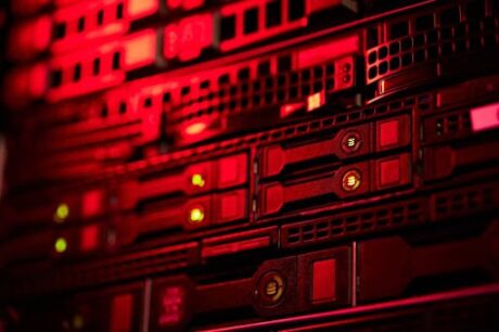 Uma fotografia horizontal de um armário de servidores iluminado por uma luz vermelha. A imagem mostra de perto várias unidades de servidores empilhadas, com luzes verdes e âmbar que indicam seu status. As luzes e a iluminação geral são dominadas pela cor vermelha. A imagem parece ser de uma sala de servidores.