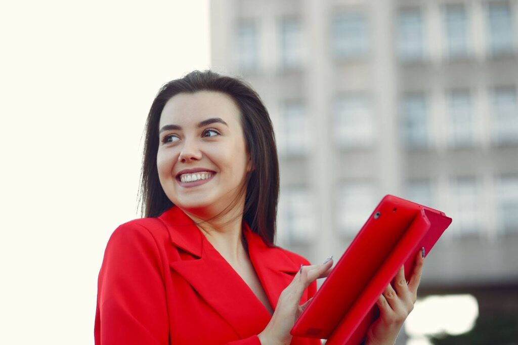 Uma fotografia horizontal de uma mulher de meia-idade, com cabelos castanhos e compridos, sorrindo e olhando para o lado. A mulher está vestindo um blazer vermelho e segurando um tablet vermelho. Ela está sorrindo e olhando para o lado, com a mão direita digitando no tablet. A imagem tem um fundo neutro, com um grande prédio embaçado em segundo plano.