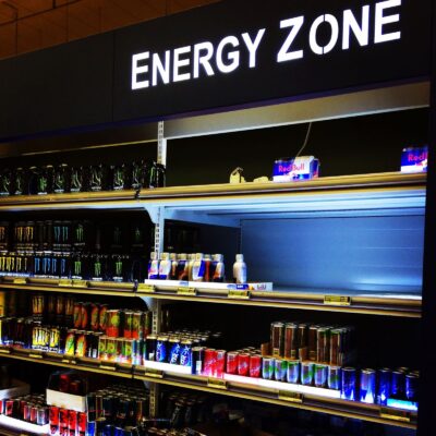 Uma fotografia de perto, horizontal, que mostra as prateleiras de uma loja. As prateleiras estão cheias de latas de bebidas energéticas, principalmente de várias marcas, como Red Bull e Monster. As latas estão em diferentes prateleiras, e algumas têm luzes azuis que as iluminam. Um letreiro grande e preto com as palavras ENERGY ZONE (ZONA DE ENERGIA) está no topo da imagem.