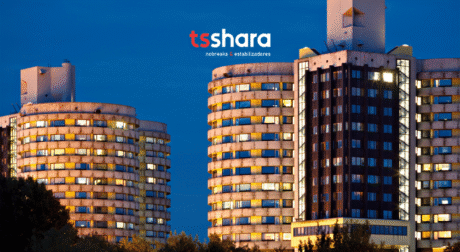 A imagem mostra dois prédios altos e modernos, com fachadas circulares, iluminados por várias janelas. O céu ao fundo está em tom azul, sugerindo que a foto foi tirada ao entardecer ou noite. O logo "tsshara" aparece no canto superior central da imagem, destacando a marca. A estrutura dos prédios é única, com um design contemporâneo e organizado.