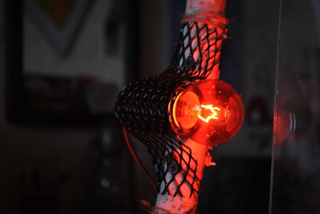 A imagem mostra uma lâmpada acesa com uma luz vermelha, envolvida por uma proteção de metal, semelhante a uma rede. O fundo é um pouco embaçado e escuro, mas há um brilho intenso vindo da lâmpada. A lâmpada parece ser de estilo antigo, com um filamento visível no centro, emitindo um brilho difuso. O ambiente ao redor da lâmpada é pouco iluminado, fazendo com que o foco principal da imagem seja a luz vermelha.