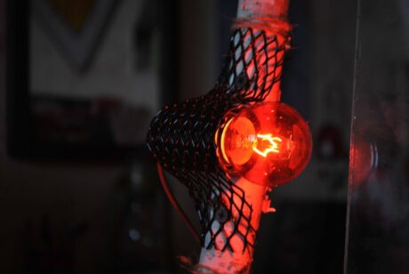 A imagem mostra uma lâmpada acesa com uma luz vermelha, envolvida por uma proteção de metal, semelhante a uma rede. O fundo é um pouco embaçado e escuro, mas há um brilho intenso vindo da lâmpada. A lâmpada parece ser de estilo antigo, com um filamento visível no centro, emitindo um brilho difuso. O ambiente ao redor da lâmpada é pouco iluminado, fazendo com que o foco principal da imagem seja a luz vermelha.