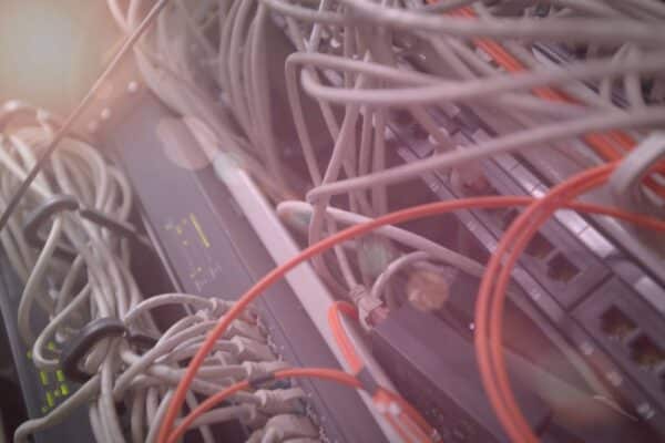A imagem mostra um close-up de cabos de rede conectados a dispositivos de um sistema de servidores. Há muitos cabos, principalmente brancos, que estão enrolados e conectados em painéis de conexão. Alguns fios são destacados em vermelho, criando um contraste visual. O fundo é ligeiramente desfocado, o que dá uma sensação de profundidade ao ambiente. A luz suave que incide sobre os cabos e os dispositivos ao fundo sugere que o local é um centro de dados ou servidor. A imagem transmite um ar de complexidade e organização técnica, com um grande número de conexões interligadas.