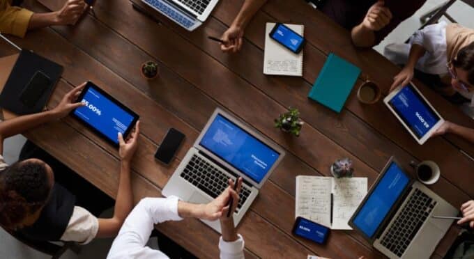 várias pessoas sentadas em uma mesa de reunião com seus aparelhos ligados, notebook, computador e celular, visto de cima