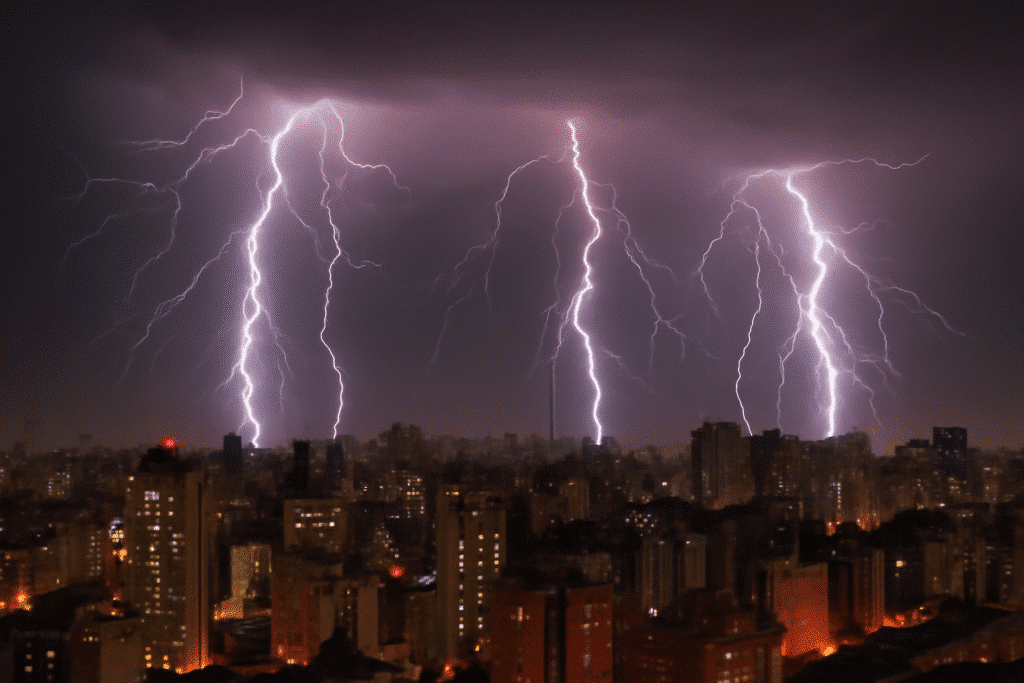 A imagem mostra uma cidade vista à noite, iluminada por raios que cortam o céu. Os raios são muito fortes e visíveis, com três deles iluminando a área central da imagem. Eles se estendem de cima para baixo, criando linhas de luz intensa, contrastando com o céu escuro. Ao fundo, há diversos edifícios urbanos, alguns com luzes acesas nas janelas, criando um efeito de cidade iluminada. A cena transmite uma sensação de energia e força, misturando a tranquilidade da cidade à fúria da tempestade no céu.