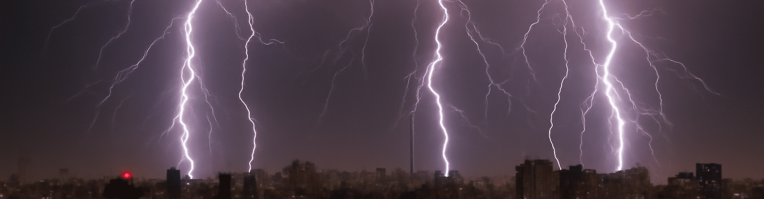 A imagem mostra uma cidade vista à noite, iluminada por raios que cortam o céu. Os raios são muito fortes e visíveis, com três deles iluminando a área central da imagem. Eles se estendem de cima para baixo, criando linhas de luz intensa, contrastando com o céu escuro. Ao fundo, há diversos edifícios urbanos, alguns com luzes acesas nas janelas, criando um efeito de cidade iluminada. A cena transmite uma sensação de energia e força, misturando a tranquilidade da cidade à fúria da tempestade no céu.