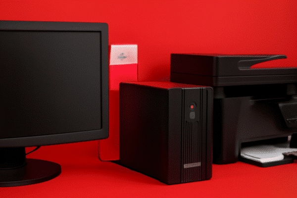 A imagem mostra uma cena com um fundo vermelho vibrante. No lado esquerdo, há um monitor de computador de tela plana, com uma moldura preta. Ao centro, há um dispositivo preto que parece ser um gabinete de computador, com uma luz amarela acesa no painel frontal. À direita, há uma impressora multifuncional preta, com uma bandeja visível onde se pode perceber folhas de papel. A composição é simples, com todos os objetos organizados de maneira que criam um ambiente de trabalho minimalista.