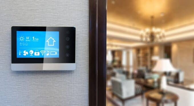 um tablet de automação ligado, preso em uma parede de uma casa e ao fundo uma sala de estar iluminada 