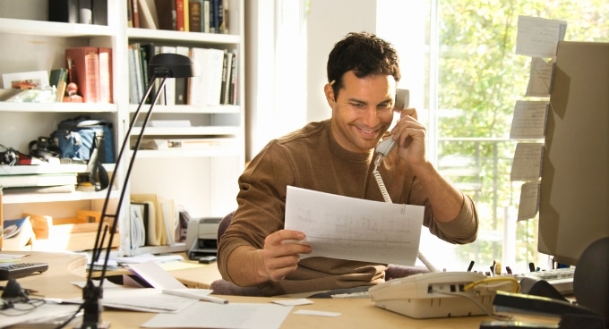 um rapaz adulto sentado falando ao telefone segurando na mão uma folha e o cenário é uma mesa com vários papéis e uma estante no fundo