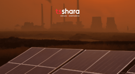 A imagem mostra, em primeiro plano, placas de energia solar em um campo aberto. Ao fundo, o céu está em tons fortes de laranja e marrom, sugerindo poluição ou fumaça intensa no ar. Na linha do horizonte, aparecem chaminés industriais e torres de resfriamento, liberando fumaça escura, remetendo a usinas ou fábricas. Na parte superior central da imagem está o logotipo da empresa "tsshara", escrito em letras minúsculas: a primeira parte "ts" em vermelho e a segunda "shara" em branco. Logo abaixo, em letras pequenas brancas, está escrito: “nobreaks e estabilizadores”. A cena contrasta a presença da poluição industrial ao fundo com a energia limpa representada pelas placas solares no primeiro plano.