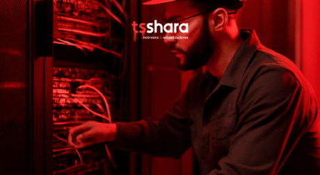A imagem mostra um profissional trabalhando em um servidor em um ambiente de data center. Ele está usando um uniforme cinza e um capacete de segurança enquanto mexe nos cabos de um equipamento, provavelmente realizando manutenção ou ajustes. A iluminação vermelha no fundo destaca o ambiente de trabalho, e o profissional parece focado em sua tarefa.