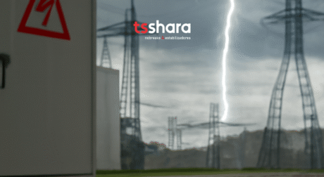 A imagem mostra um cenário externo de torres de energia elétrica altas, em meio a um céu cinzento e carregado. Um raio branco intenso corta o céu ao fundo, chamando bastante atenção. No lado esquerdo da imagem, aparece parcialmente uma estrutura branca com um símbolo vermelho de alerta elétrico. Na parte superior central da imagem está o logotipo da marca “tsshara”, escrito em letras minúsculas: o “ts” em vermelho e o “shara” em branco. Logo abaixo, em letras menores e brancas, lê-se: “nobreaks e estabilizadores”. A cena transmite a ideia de risco elétrico e a importância de proteção contra quedas ou surtos de energia.