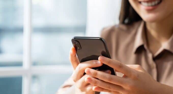 As mãos de uma mulher segurando um smartphone preto na vertical, com o foco na tela e na parte traseira superior onde fica a câmera.