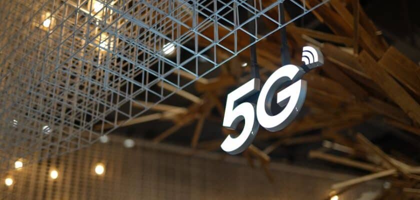 Um letreiro luminoso com o símbolo "5G" está pendurado no teto, iluminado em branco, com um ícone de sinal Wi-Fi ou de rede sobre o "G"