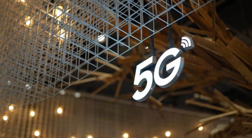 Um letreiro luminoso com o símbolo "5G" está pendurado no teto, iluminado em branco, com um ícone de sinal Wi-Fi ou de rede sobre o "G"