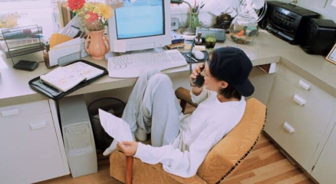 uma mulher sentada na poltrona de casa em frente ao computador trabalhando e falando ao telefone