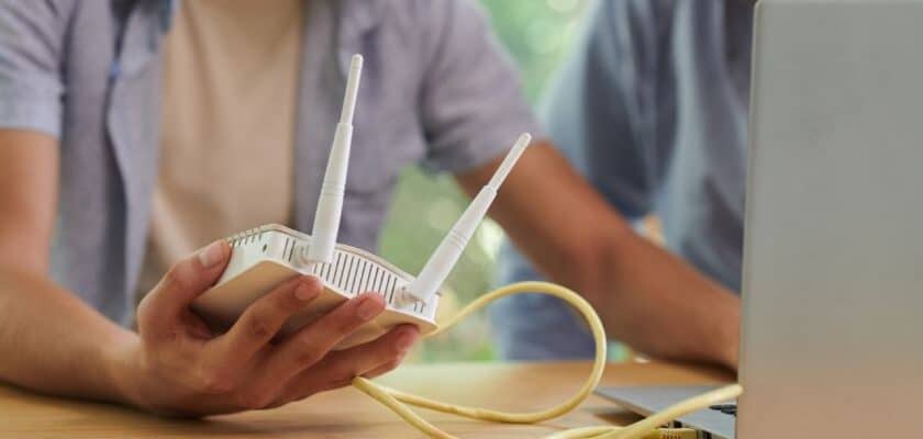 uma mão de um rapaz segurando um roteador de internet na mão, enquanto sua outra mão está em um notebook tentando conectar