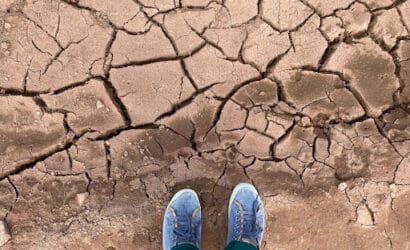 Pés calçados com tênis azuis sobre solo seco e rachado, indicando falta de umidade.
