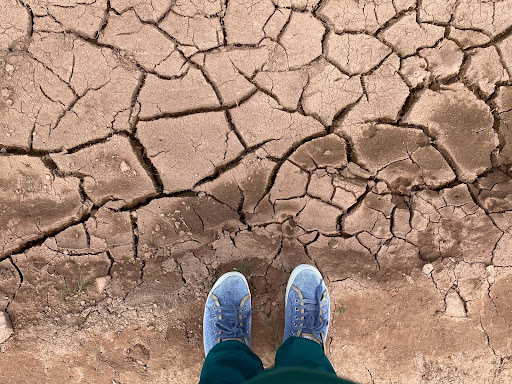 Pés calçados com tênis azuis sobre solo seco e rachado, indicando falta de umidade.