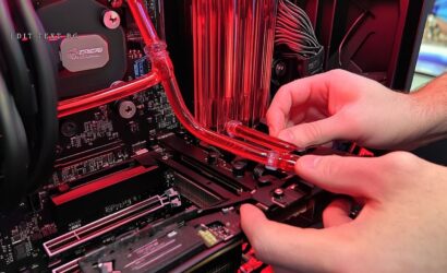 A imagem mostra o processo de montagem de um sistema de resfriamento a água para um computador. É possível ver tubos vermelhos conectando o sistema, enquanto uma mão está ajustando os tubos na parte interna do gabinete. O fundo da imagem tem iluminação vermelha, destacando o hardware e os componentes, como a placa-mãe e a placa gráfica. A estética da imagem é moderna e técnica, destacando a construção de um PC customizado.